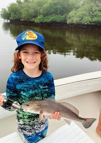 A smiling young boy wearing a Bass Pro Shops hat holds up a redfish on a boat; a water and wooded shoreline is in the background.