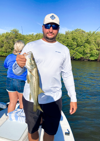 Man holding a fish on a boat, sunny day with mangroves in background. He is wearing a hat and sunglasses.