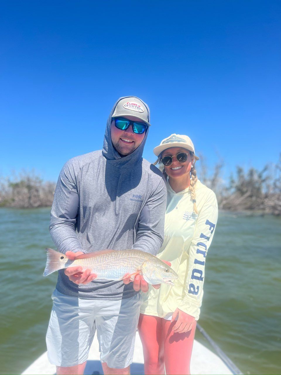 A man and woman on a boat hold up a redfish they caught in Florida waters under a blue sky. They are smiling.