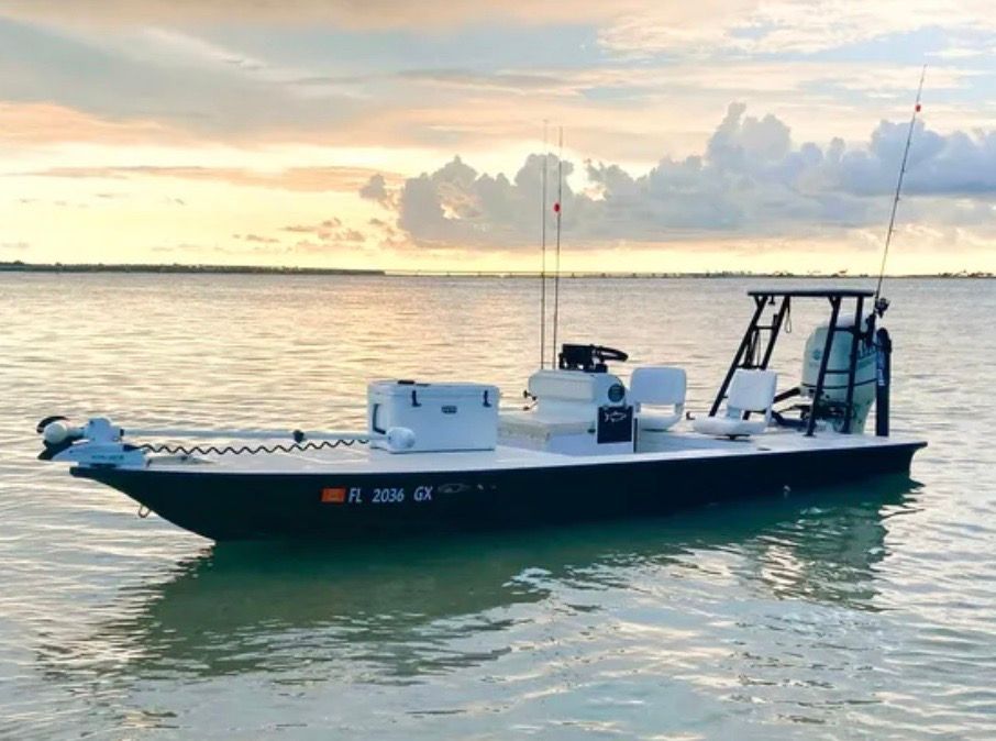 A flat bottom fishing boat with a dark hull floats in calm water under a cloudy sky.