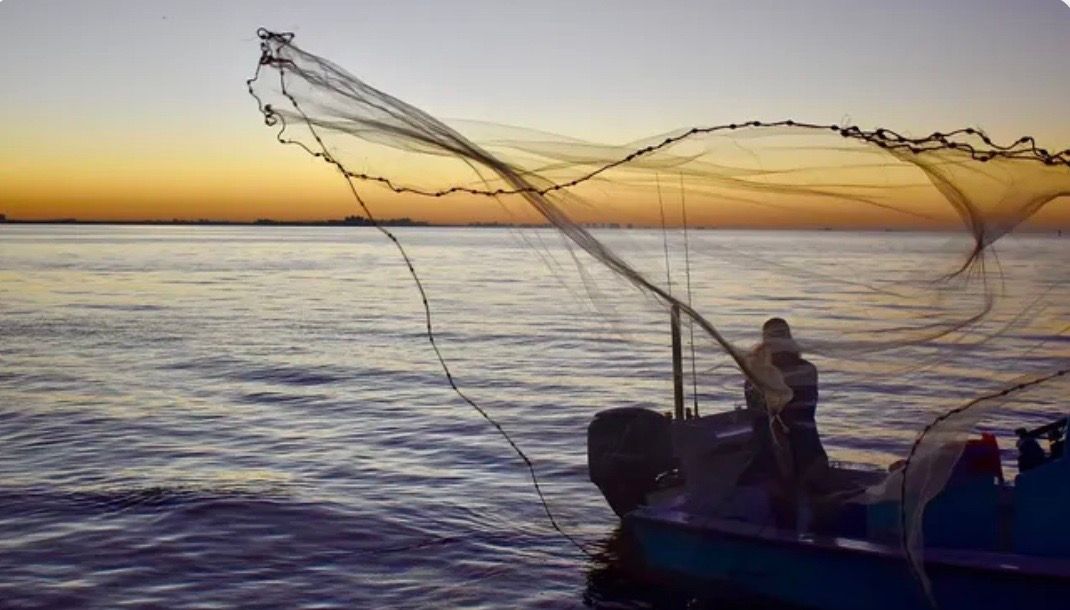 Fisherman casting a net from a boat on the water at sunset.