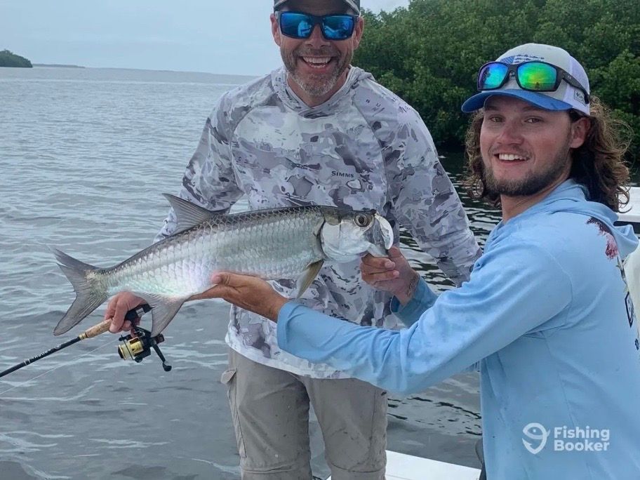 Two men on a boat display a large silver fish they caught; one man holds it, smiling, against a cloudy sky.