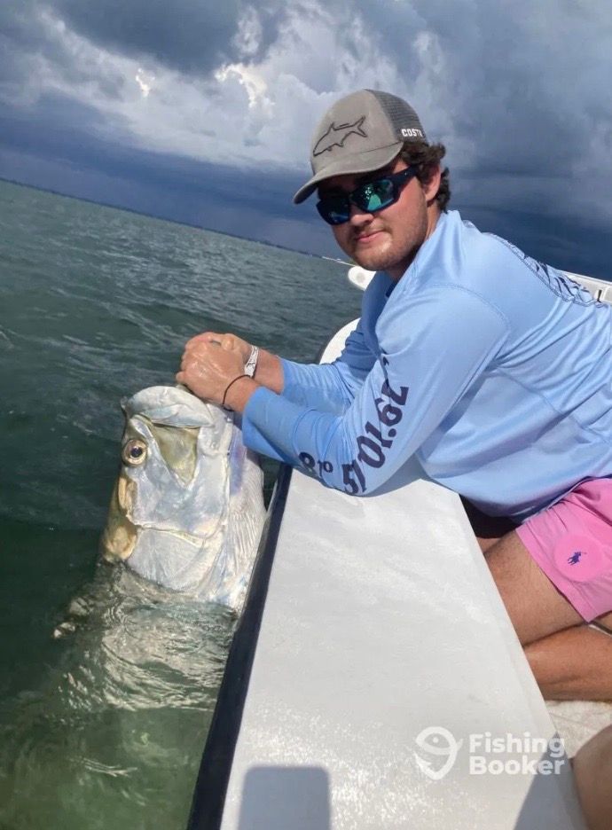 A person on a boat holds a large tarpon in the water. The person wears sunglasses and a hat, and the sky is cloudy.