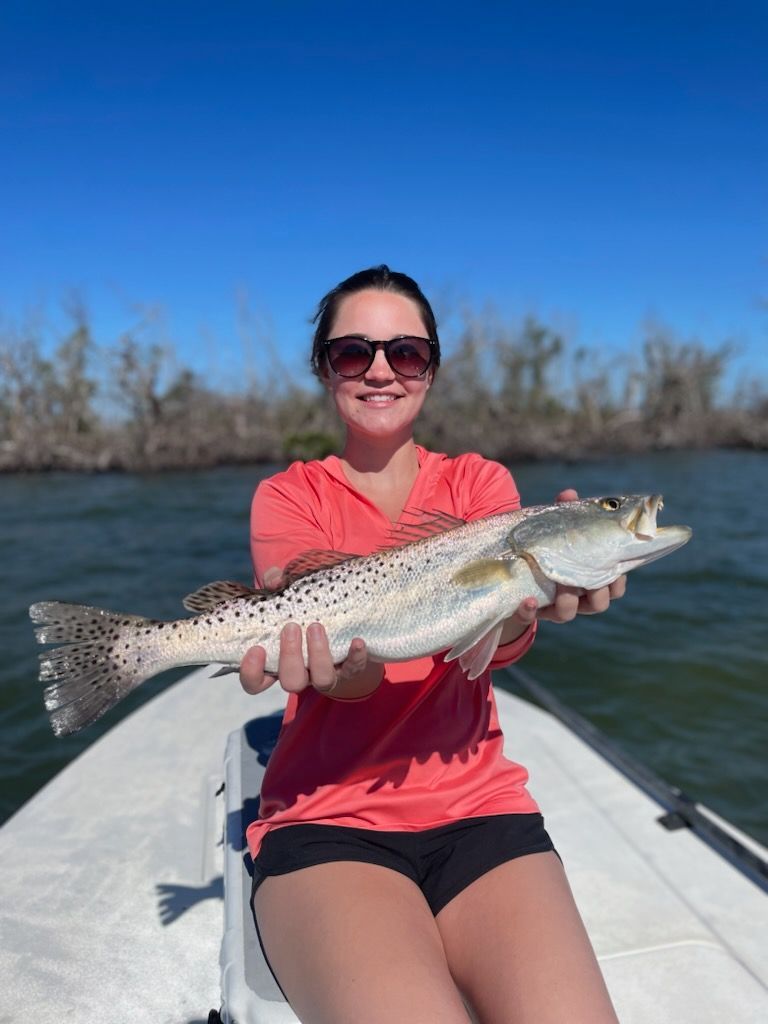 Woman on a boat holds up a speckled trout, smiling. She wears a pink shirt and sunglasses; blue sky and water in the background.