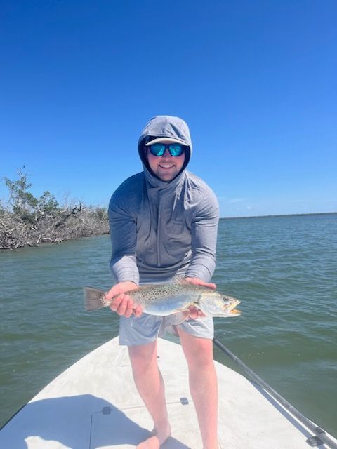 Man on a boat holds a speckled trout; clear blue sky, sun, and coastal landscape in the background.