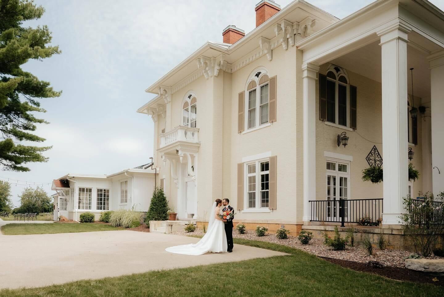 A wedding couple stands together in front of a large, historic cream-colored mansion with white pillars and trim.