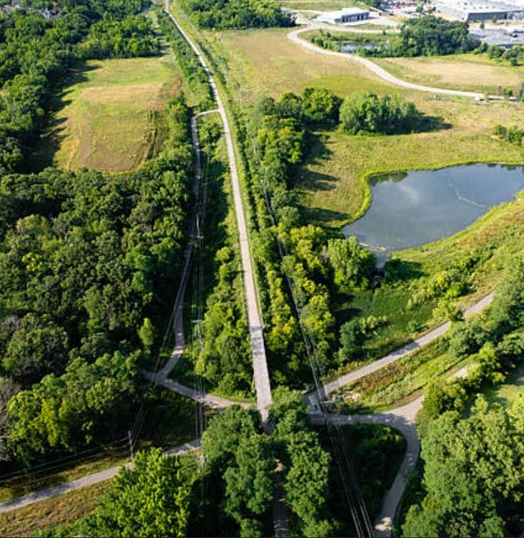 An aerial view shows a straight, paved trail running through a green landscape with trees, a small pond, and nearby fields.