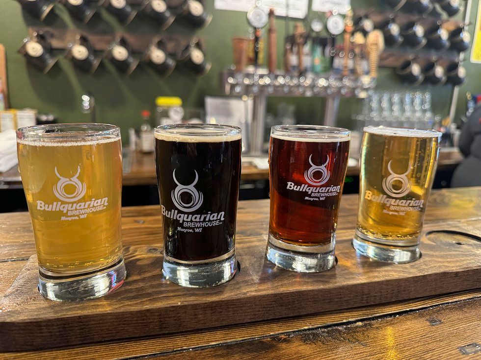 A beer flight with four glasses of varying shades of beer on a wooden board at a brewery bar.