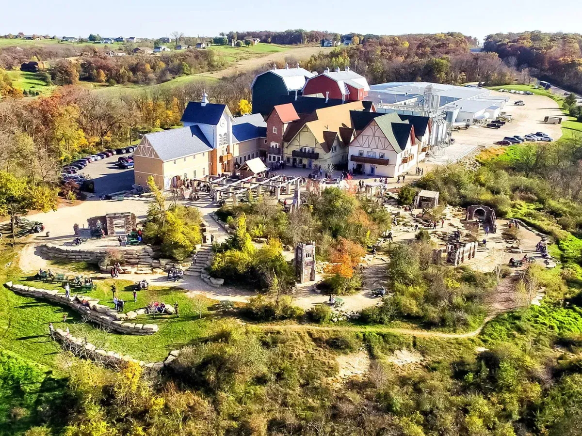 An aerial view of a rustic, multi-building complex nestled in a forested landscape, featuring stone pathways and ruins.