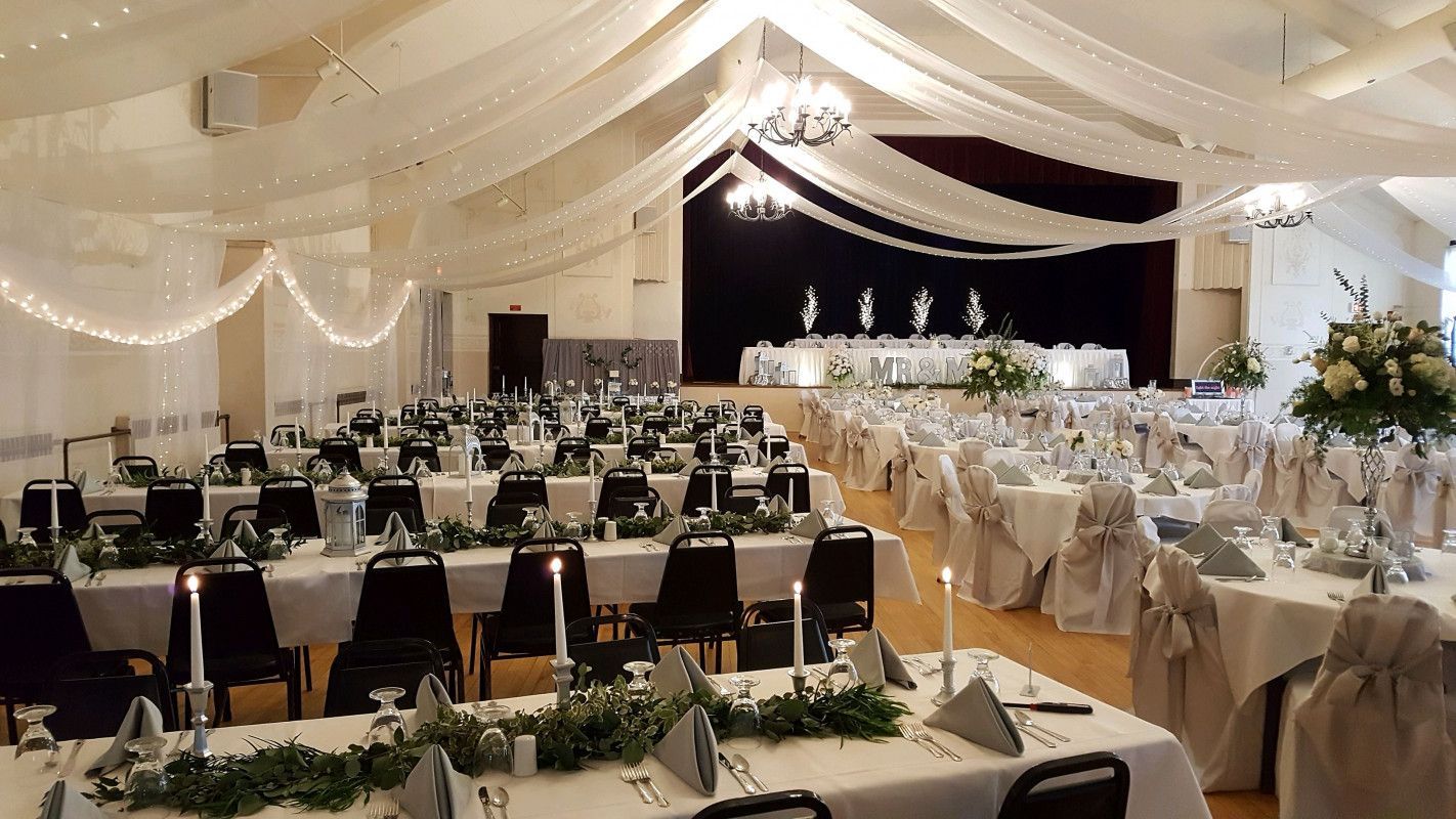 A spacious event hall decorated for a formal reception with white linens, floral centerpieces, and white ceiling draping.