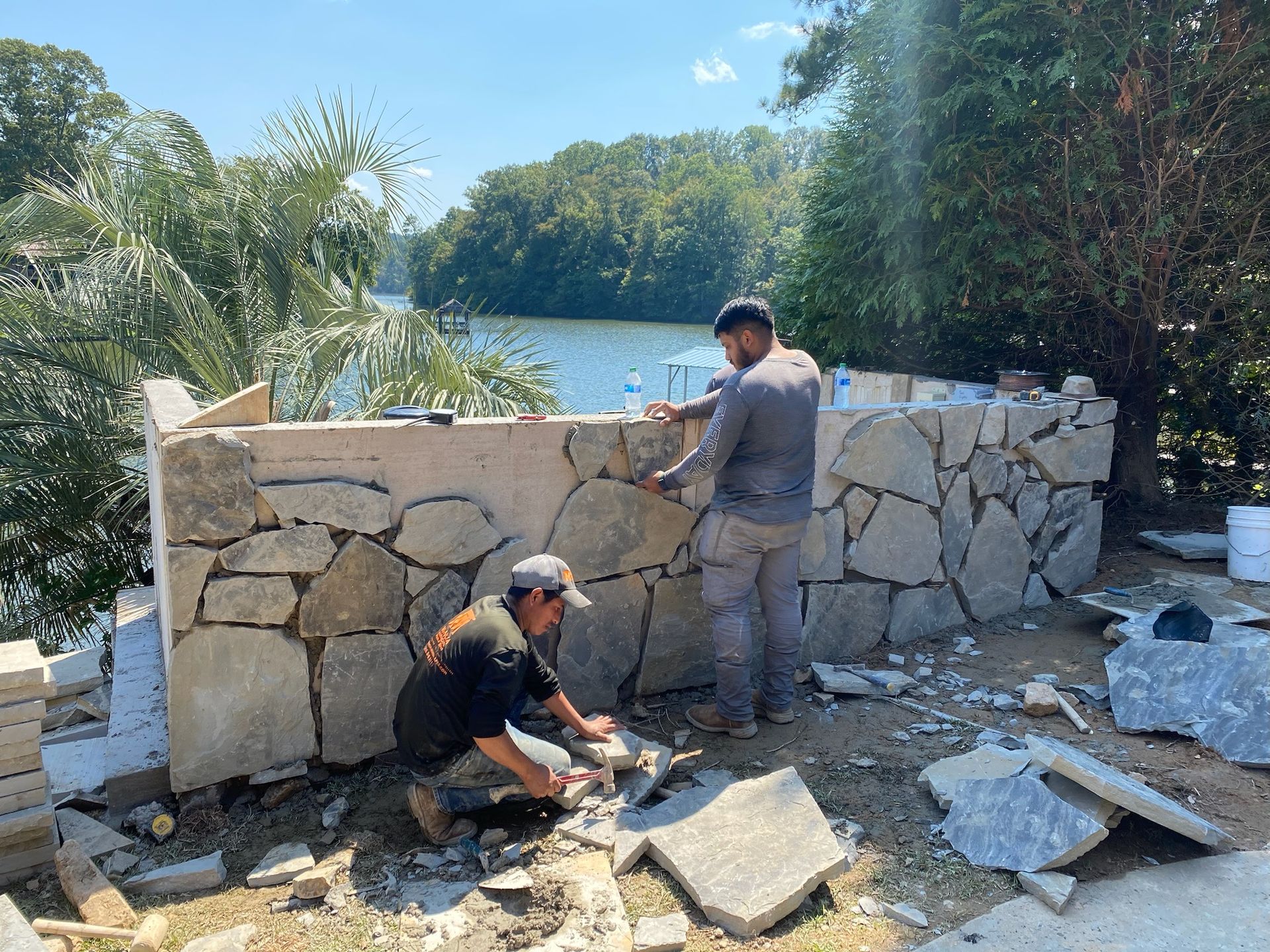 Two men are working on a stone wall in front of a lake.