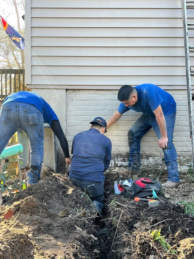 Three workers in blue shirts and jeans kneel in the dirt, excavating along the foundation of a house with beige siding.