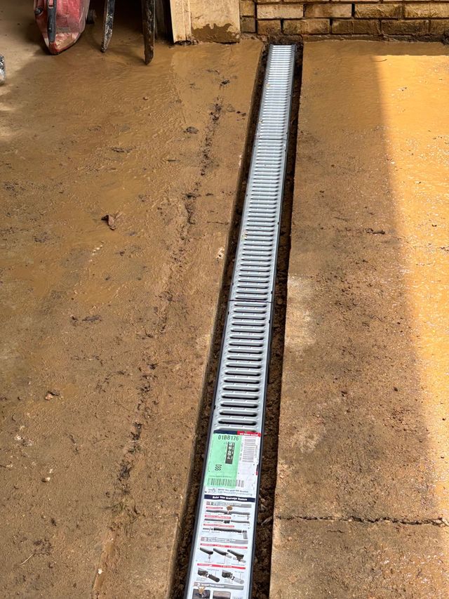 A silver plastic drainage channel installed in a concrete walkway, with a product label visible on the top section.