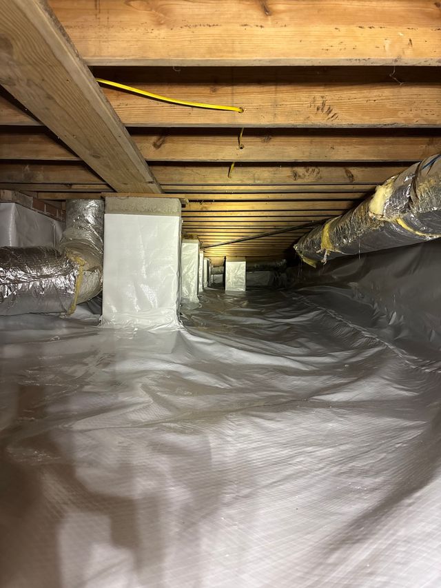Crawl space with white vapor barrier, metal support piers, and silver insulated ductwork under wooden floor joists.