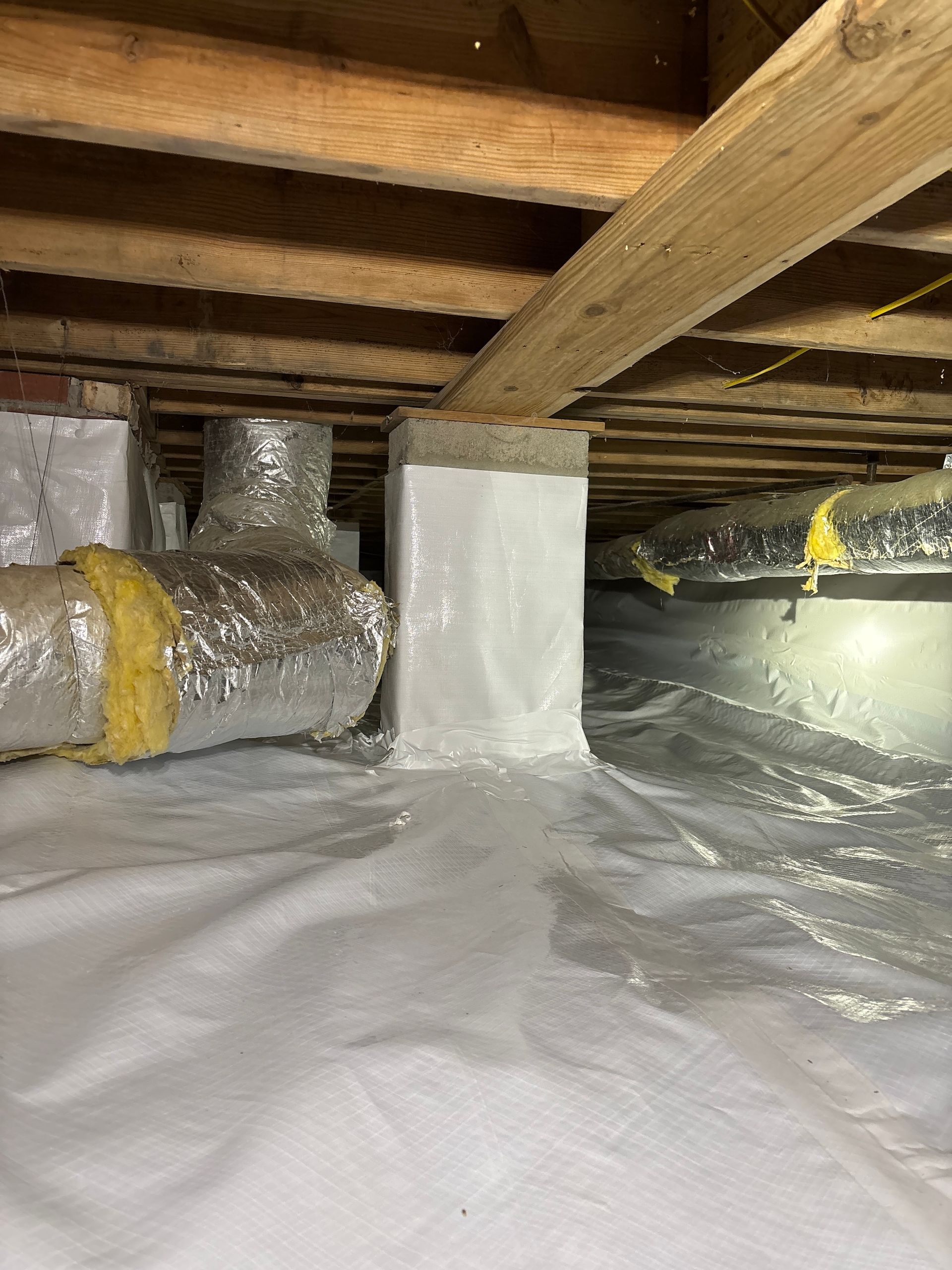 A crawl space with insulated air ducts and a white vapor barrier covering the ground and a concrete support pier.
