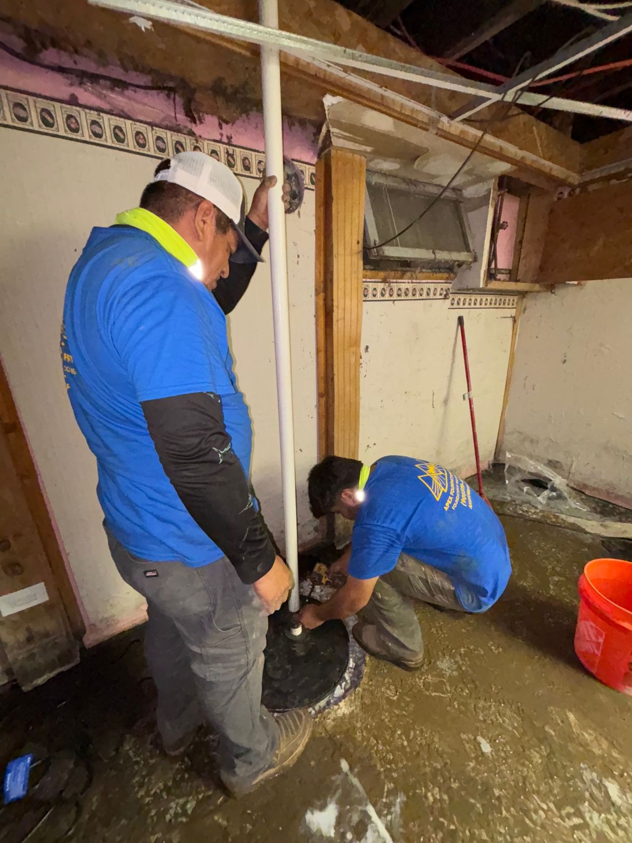 Two workers in blue shirts install a vertical pipe into a hole in a concrete floor in an unfinished basement.