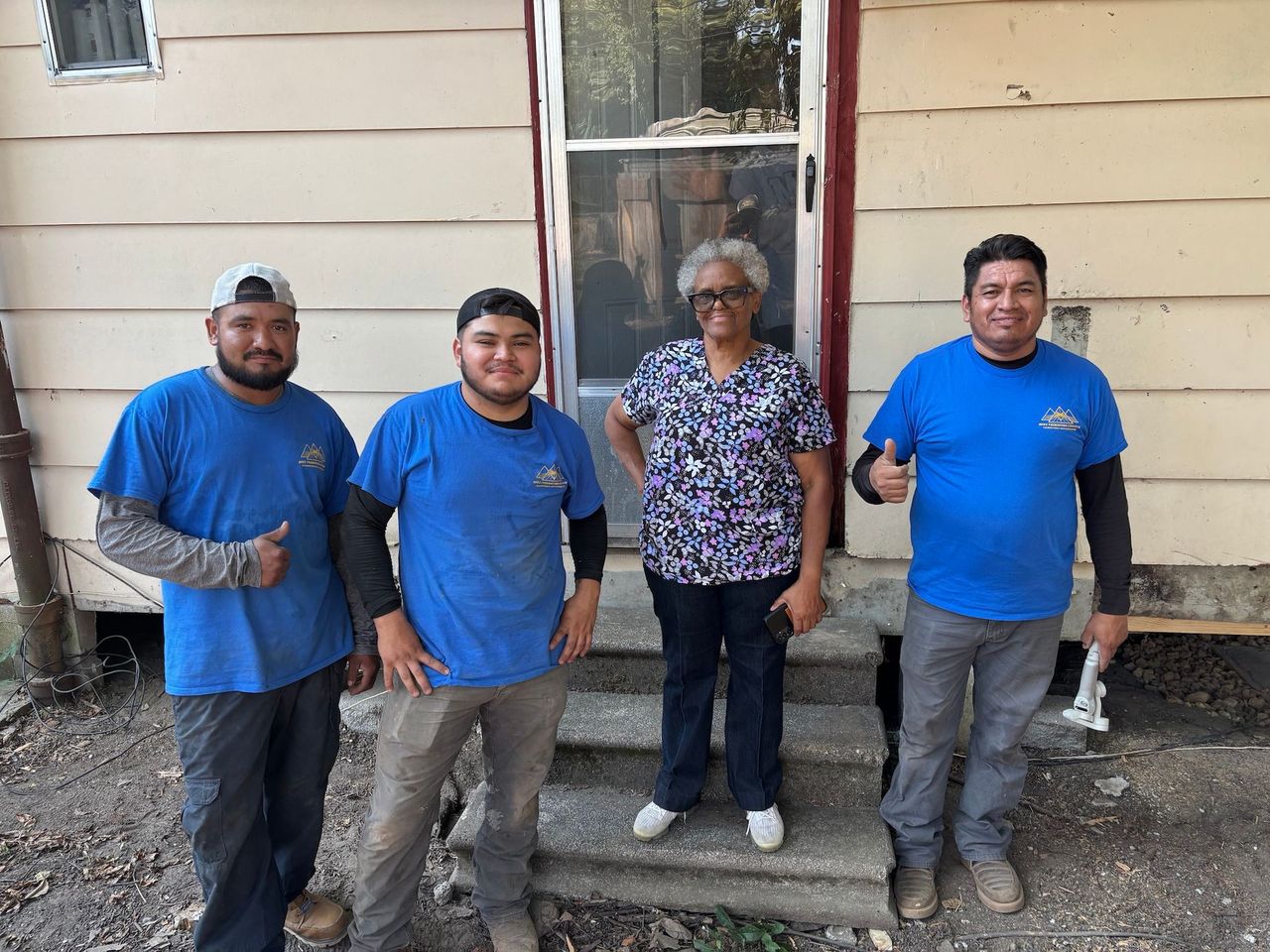 Four people in work clothes standing in front of a house, with three people giving a thumbs-up gesture.