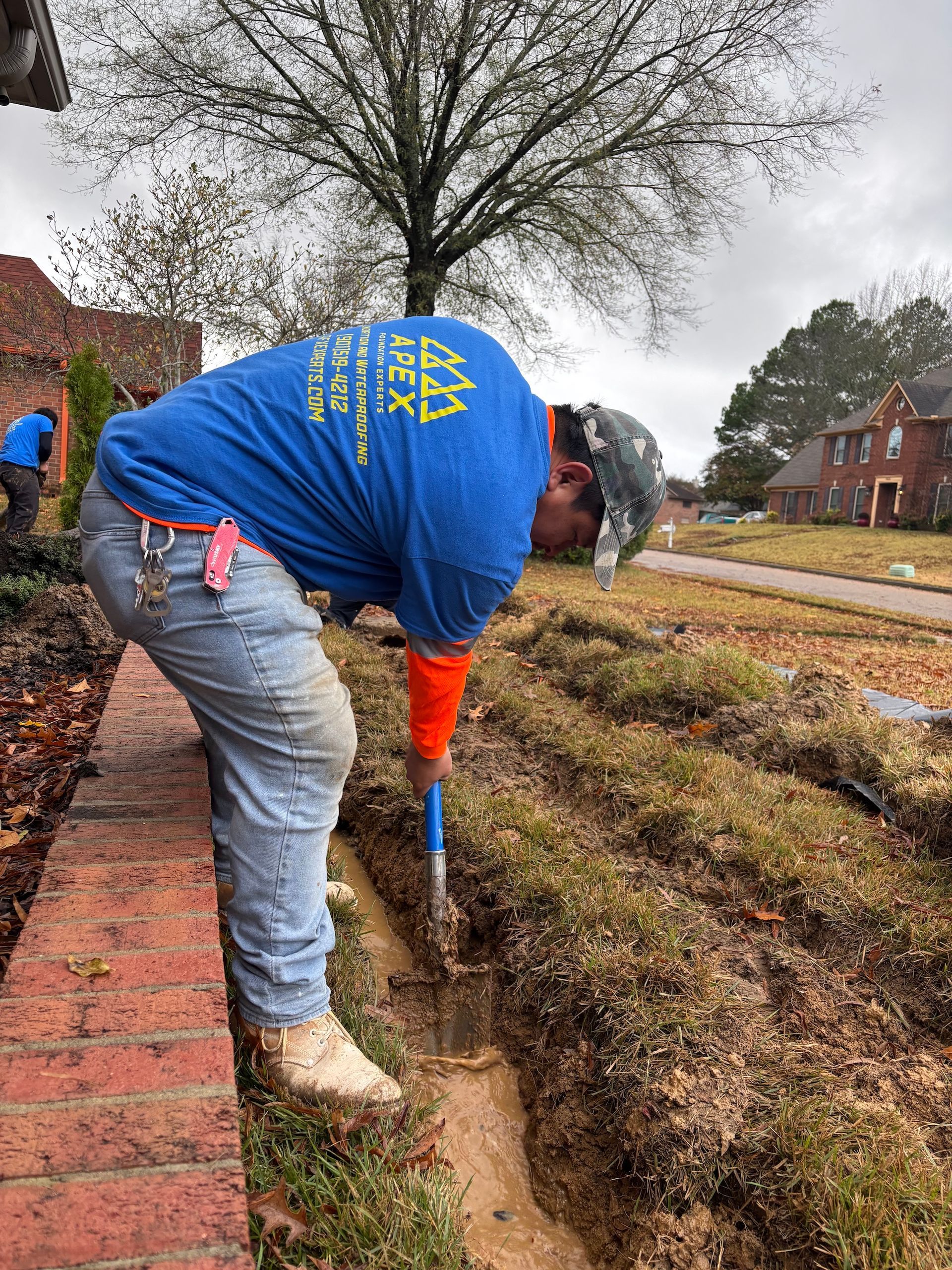 A worker in a blue t-shirt digs a trench along a brick retaining wall in a grassy front yard.