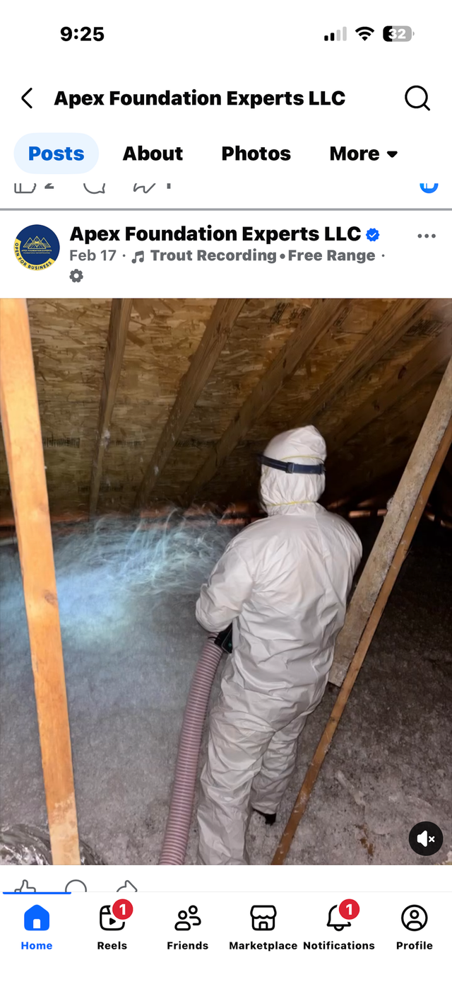A worker in a white protective suit uses a hose to spray insulation in an attic.