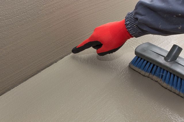 A gloved finger points to the corner where a freshly troweled concrete floor meets a wall next to a scrubbing brush.