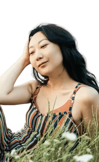 Woman in striped jumpsuit, hand in hair, sitting in tall grass, looking relaxed.