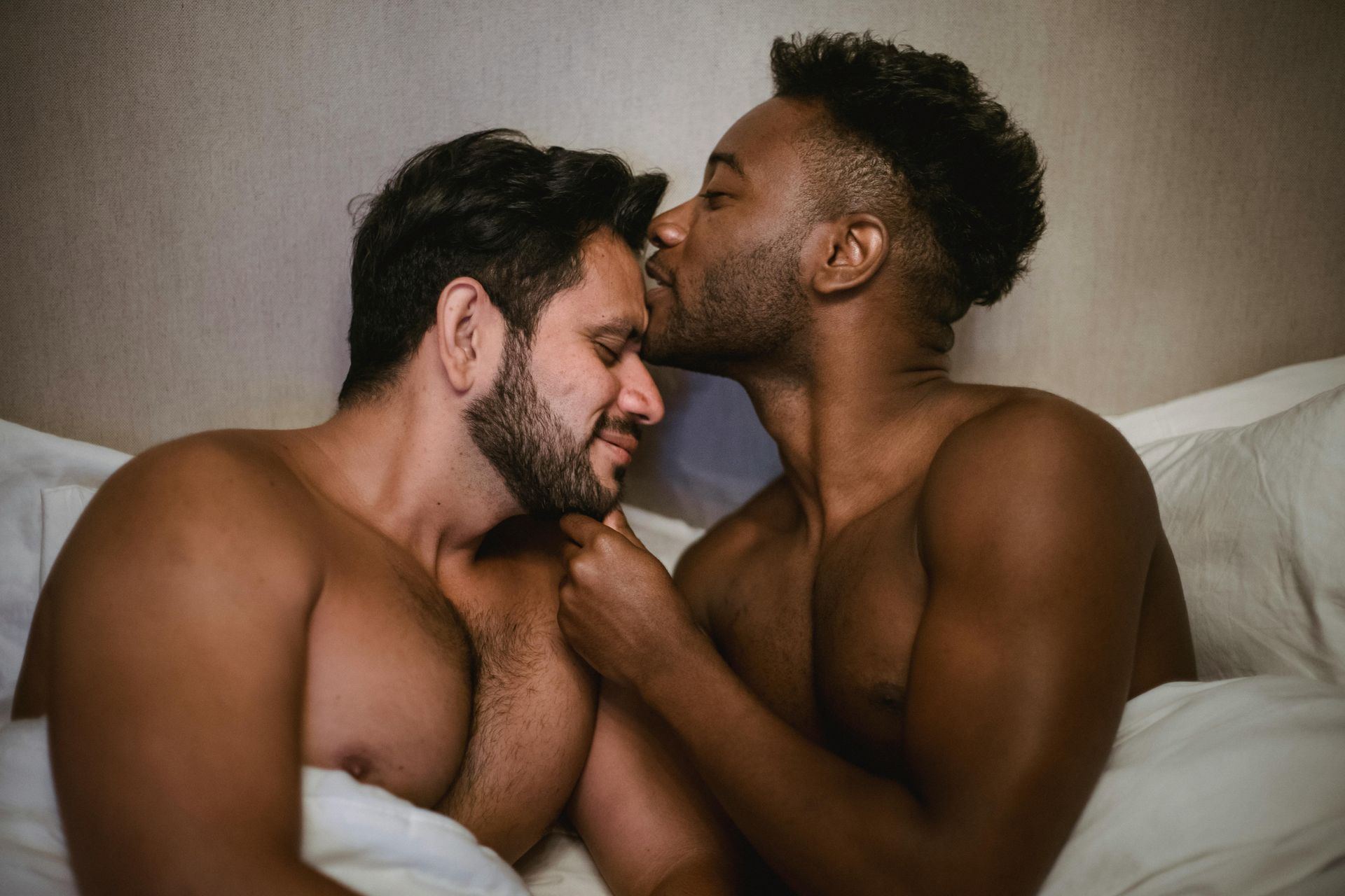Two men in bed, one kissing the other's forehead.