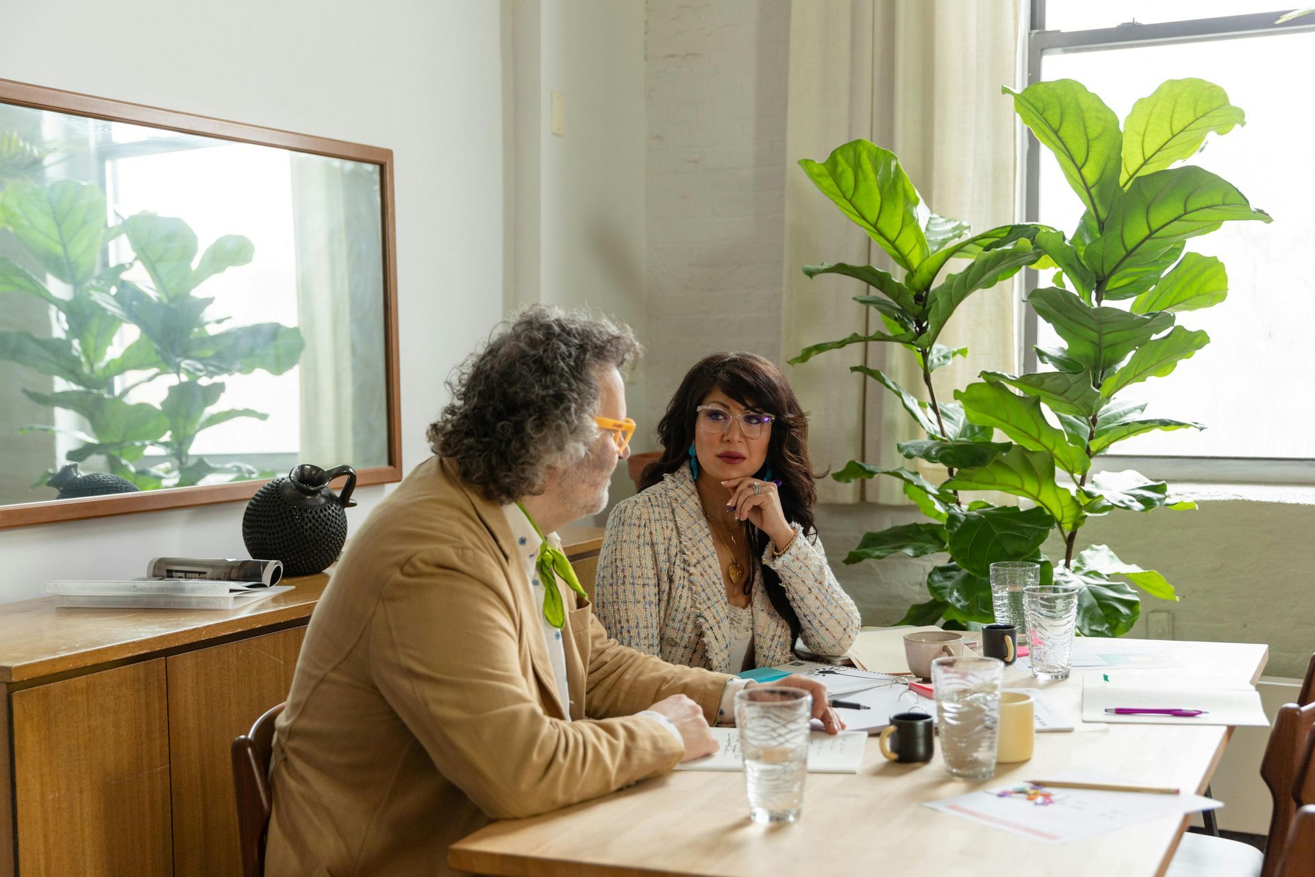 Two people sit at a table in a bright, modern office with a large plant, reviewing documents during a meeting.