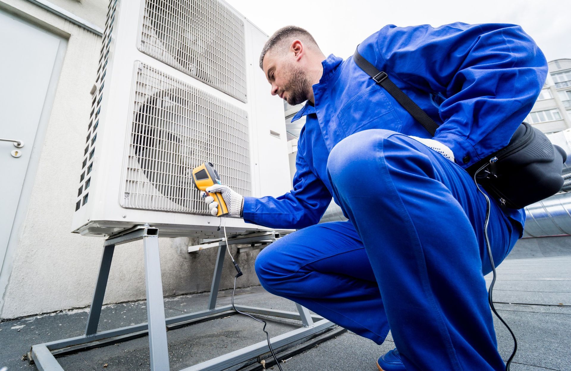 A man in blue overalls is kneeling down and looking at an air conditioner.