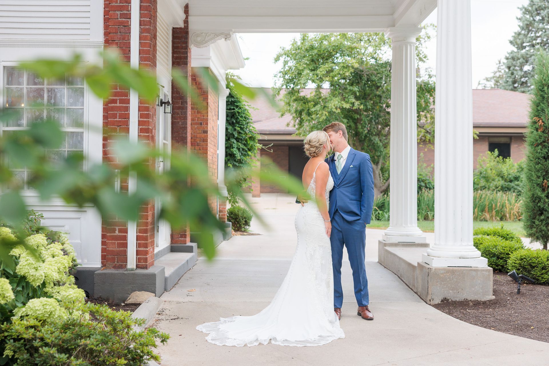 A bride and groom are kissing under a porch in front of a brick building.