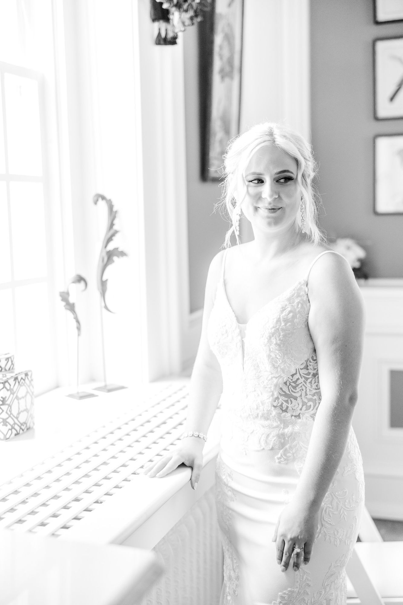 A black and white photo of a bride in a wedding dress standing next to a window.