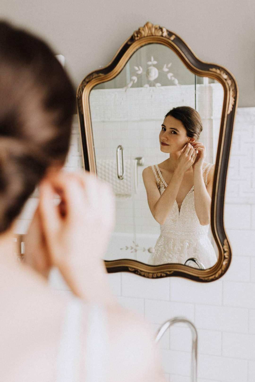 A woman in a wedding dress is putting on her earrings in front of a mirror.