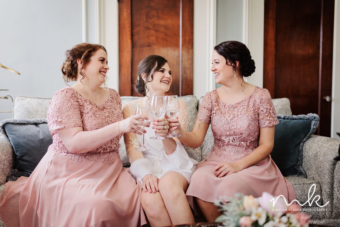 A bride and her bridesmaids are sitting on a couch toasting with wine glasses.