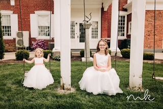 Two flower girls are sitting on swings in front of a brick building.