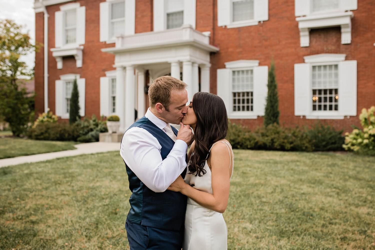 A bride and groom are kissing in front of a large brick building.