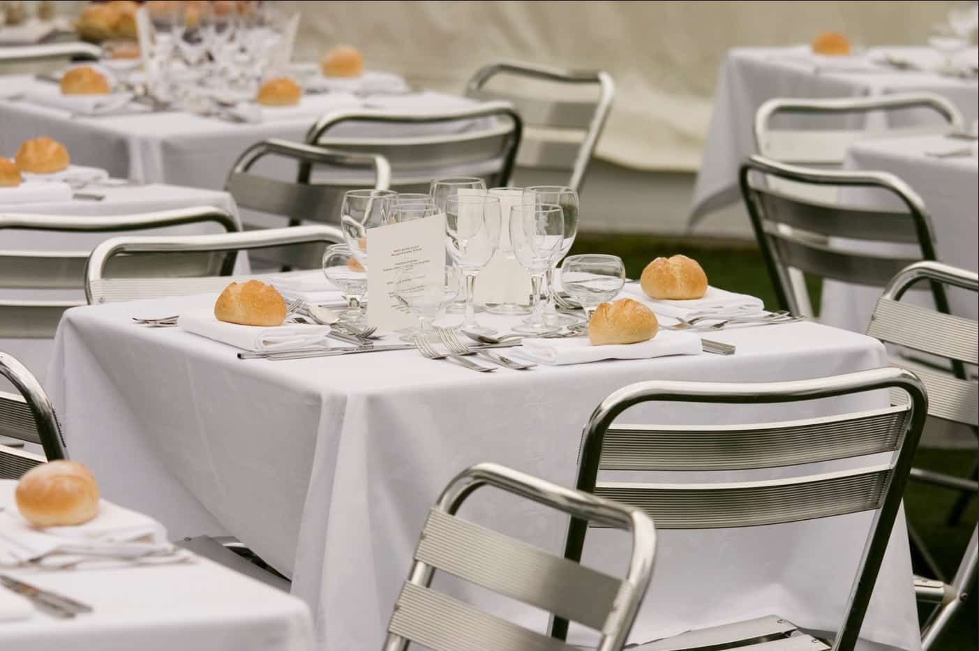 Tables set with white tablecloths, silverware, bread rolls, and glasses, outdoors.