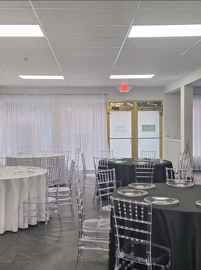 Event room with round tables, chairs, and sheer curtains. Black and white tablecloths, clear chairs.