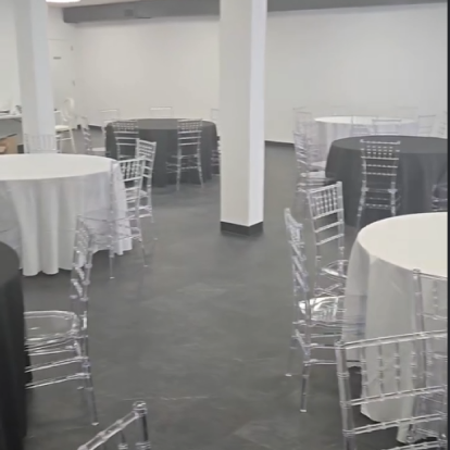 Empty event space with round tables in white and black tablecloths, clear chairs.