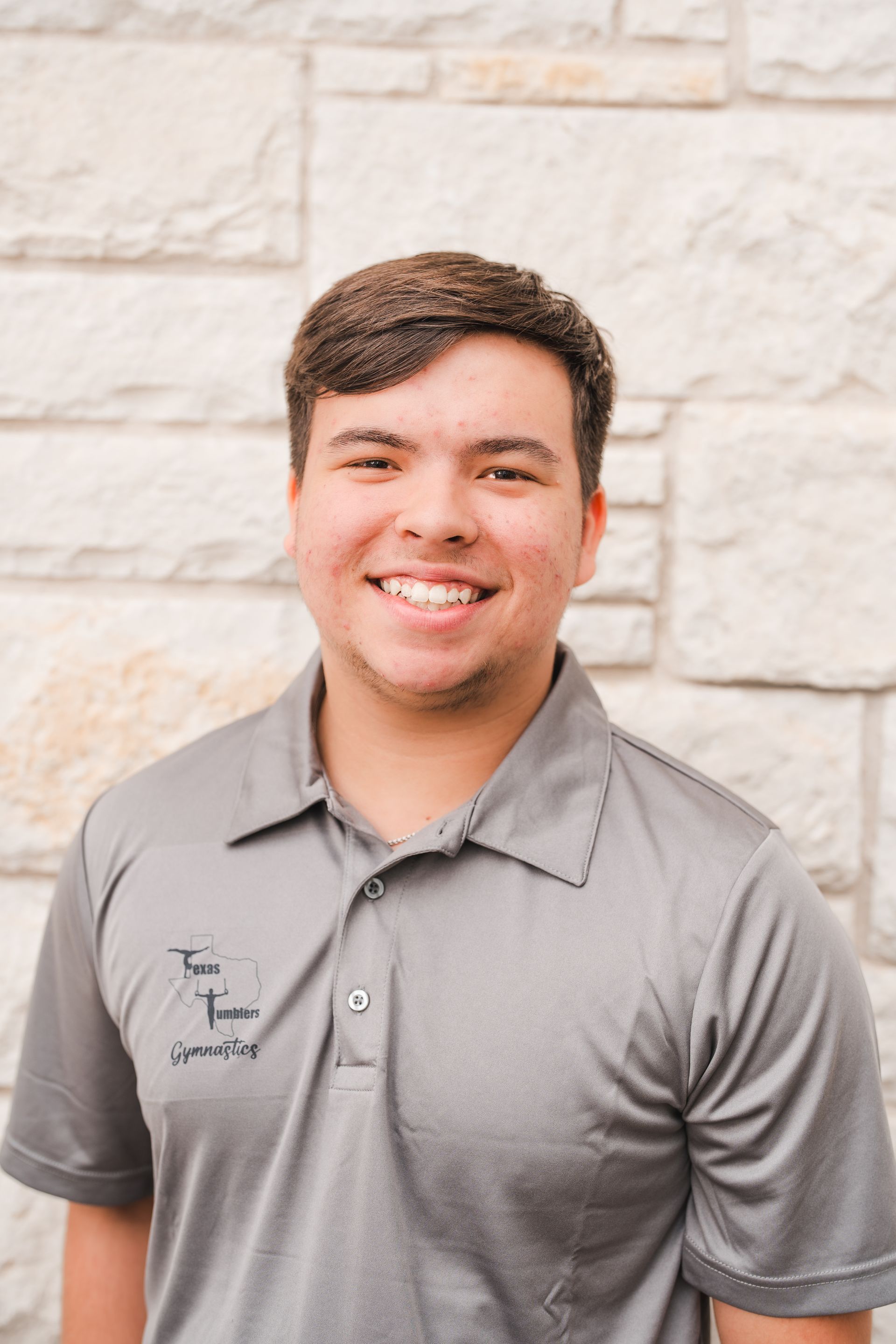 A young man in a grey polo shirt is smiling in front of a brick wall.