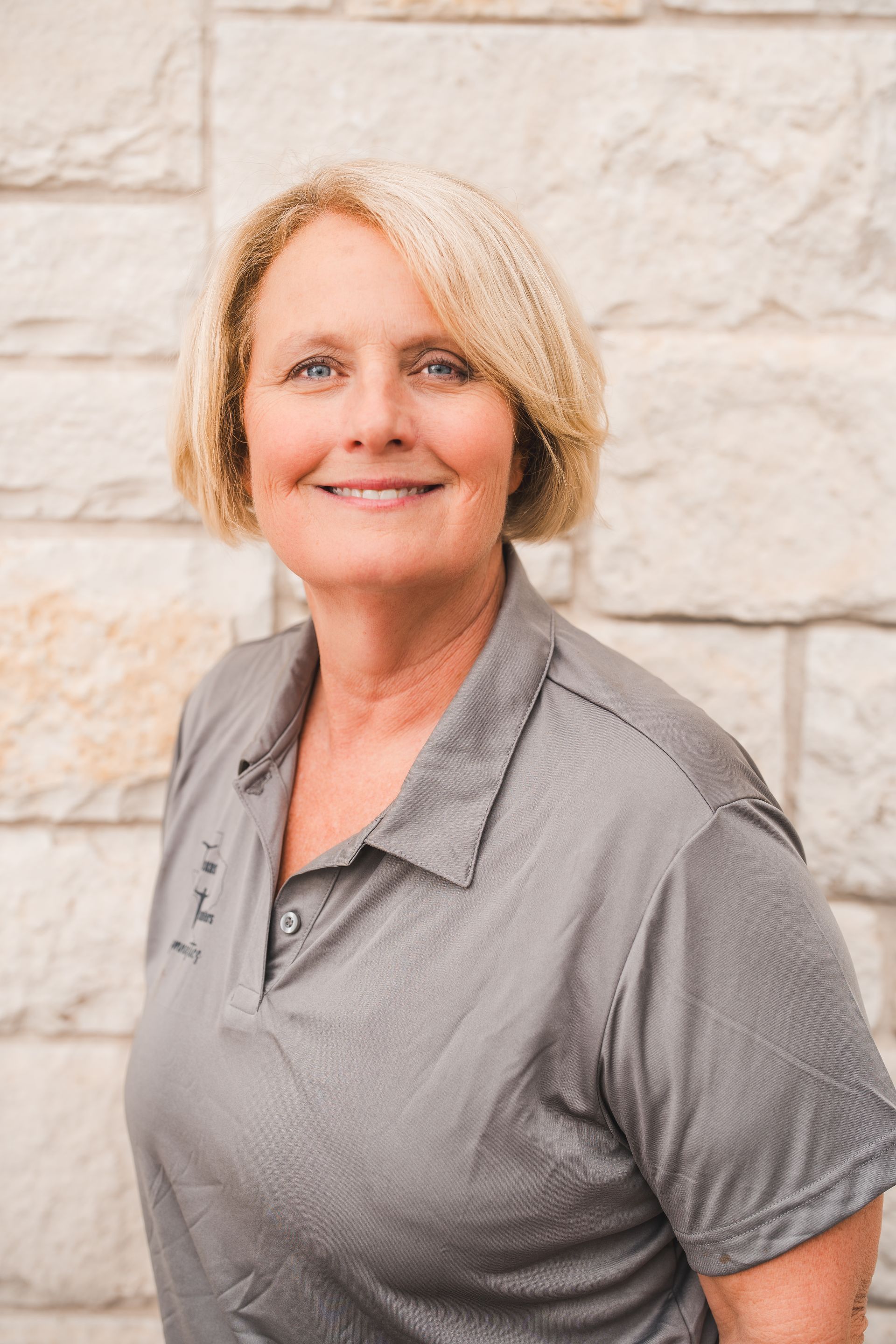 A woman in a grey shirt is smiling in front of a brick wall.