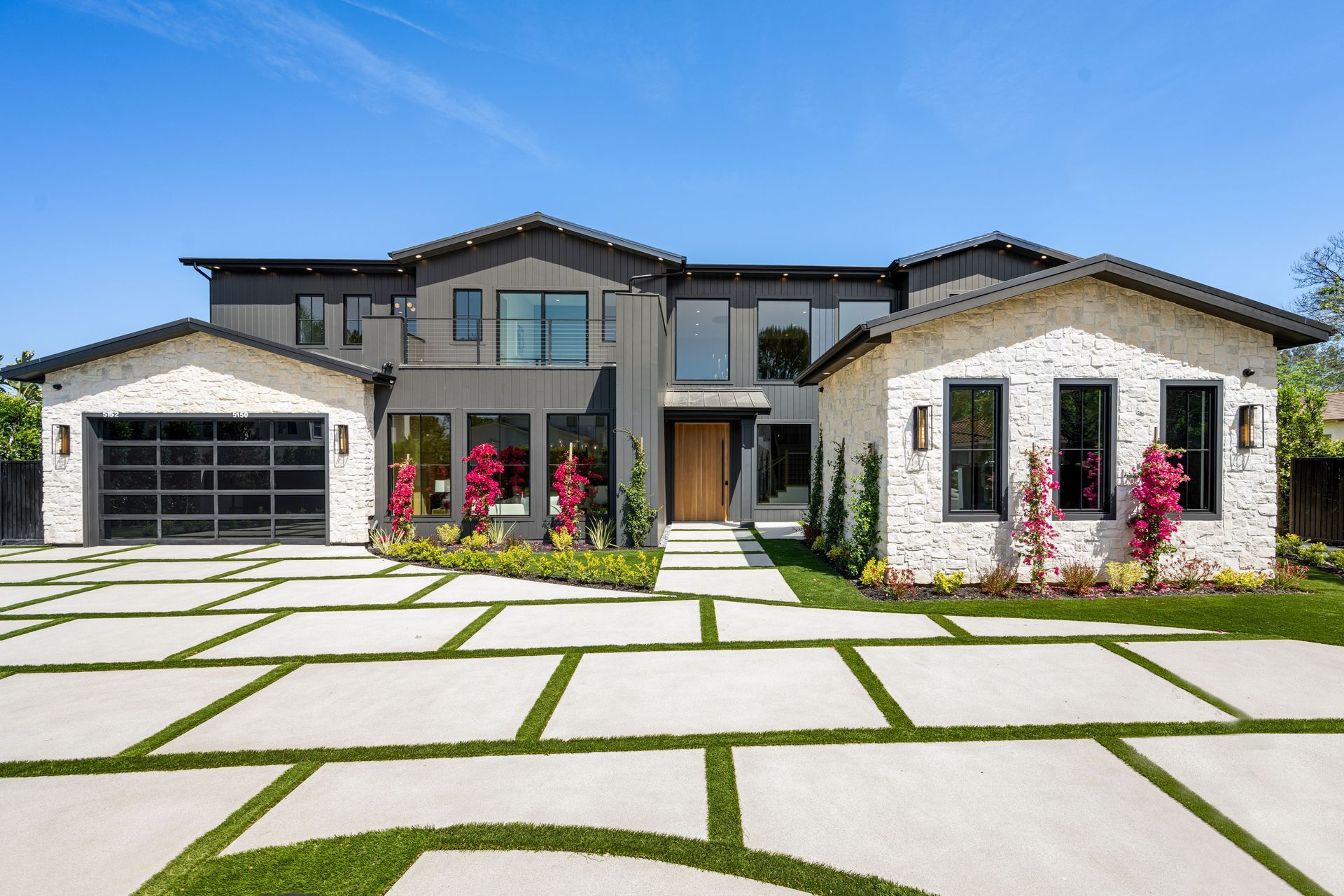 Modern two-story house with gray and white exterior. Wide driveway with square stepping stones and green grass.
