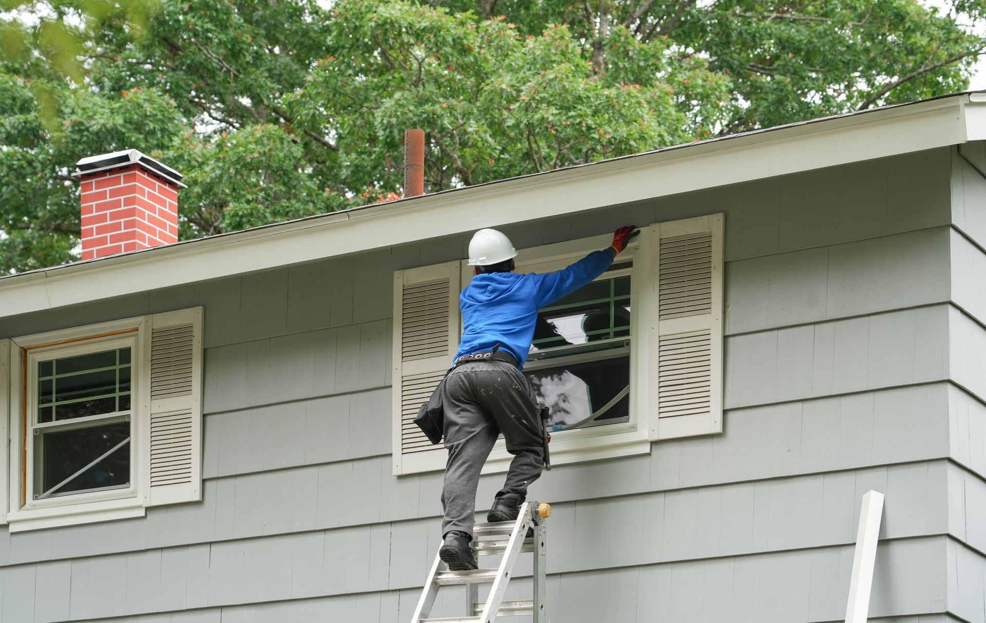Person on ladder repairing house window, wearing hard hat.