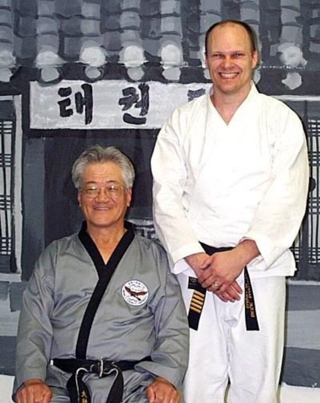 Two men are posing for a picture in front of a sign that says tae kwon do