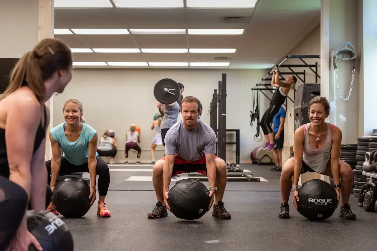 People in a gym performing wall ball exercises. Some others are doing pull-ups and lifting weights.