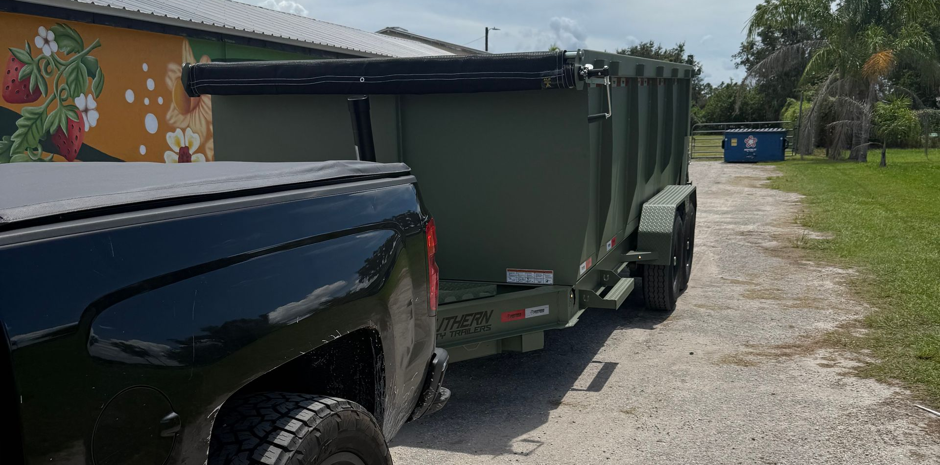 Black truck towing a green dumpster trailer on a gravel road. Mural on a wall in the background.