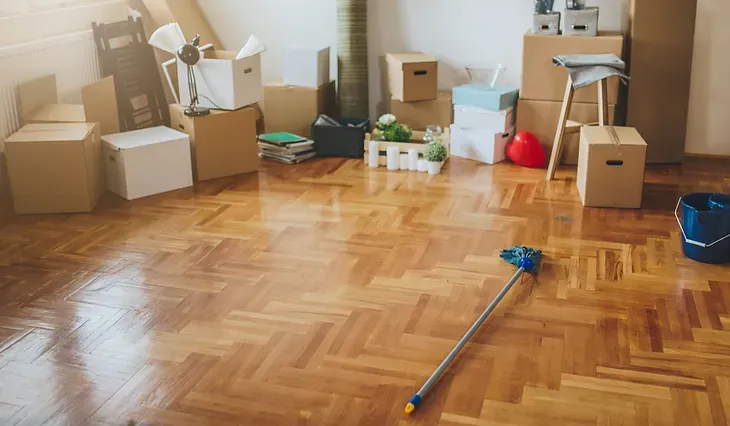 A room with packed boxes and a mop. Brown hardwood floor.