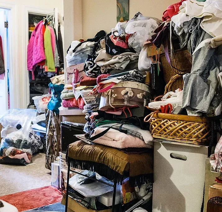 A cluttered room with piles of clothing and objects stacked high. An open closet with hanging clothes is visible.