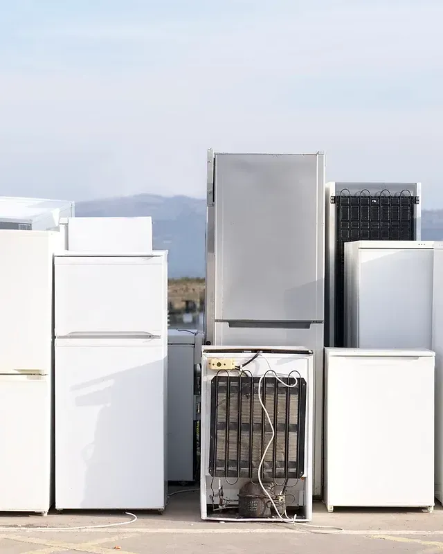 Old refrigerators on a flat surface, some white, one with back exposed. Sky in background.