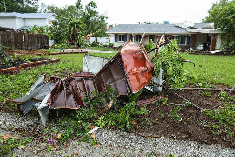 Damaged metal shed and debris in front of a house on a grassy lawn after a storm.