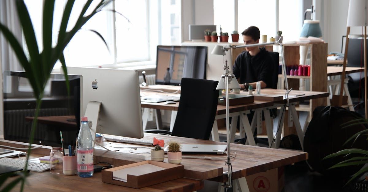 Man working at a desk with computer, plants, and supplies in a bright office space.