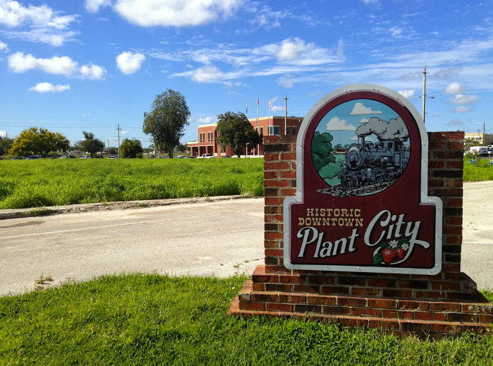 Sign for Historic Downtown Plant City with a train illustration, field, and building under a blue sky.